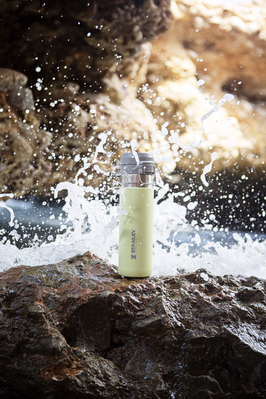A green Stanley bottle on coastal rocks surrounded by a breaking wave